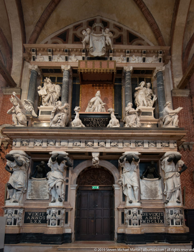 Large multi-tiered funerary monument fills a brick wall, with many marble figures, dark inscriptions, heraldry, and a central doorway.