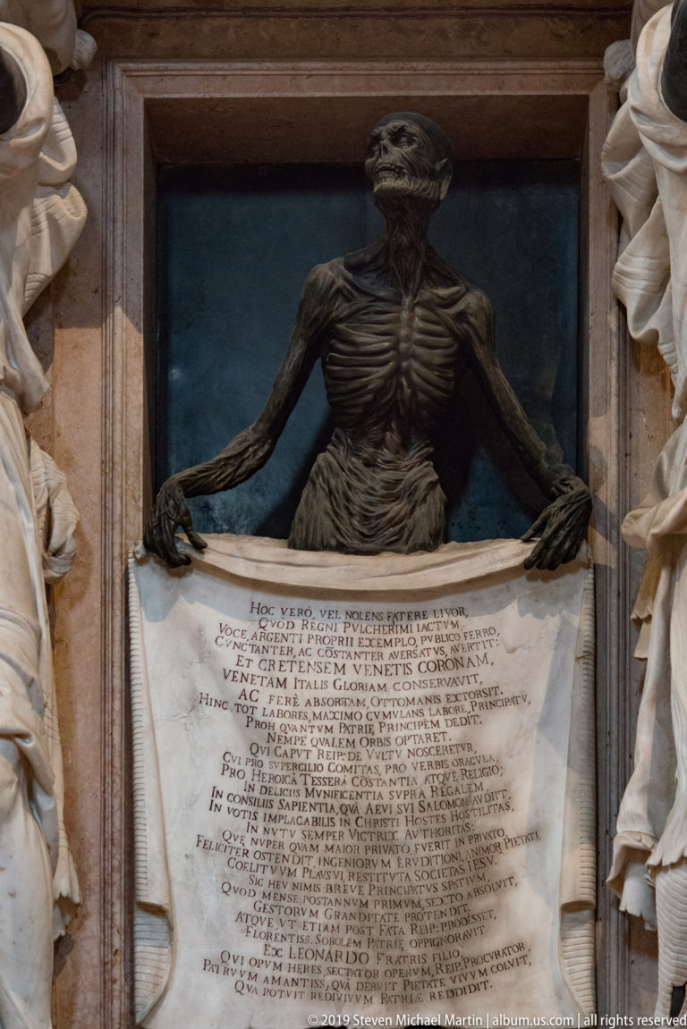 Skeletal dark figure with exposed ribs holds the top corners of a large Latin funerary inscription, with pale draped figures at the edges.
