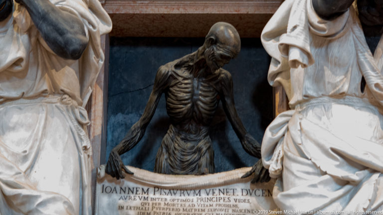 Close view of a dark skeletal figure with bowed head and exposed ribs gripping an inscribed funerary panel between pale draped sculptures.