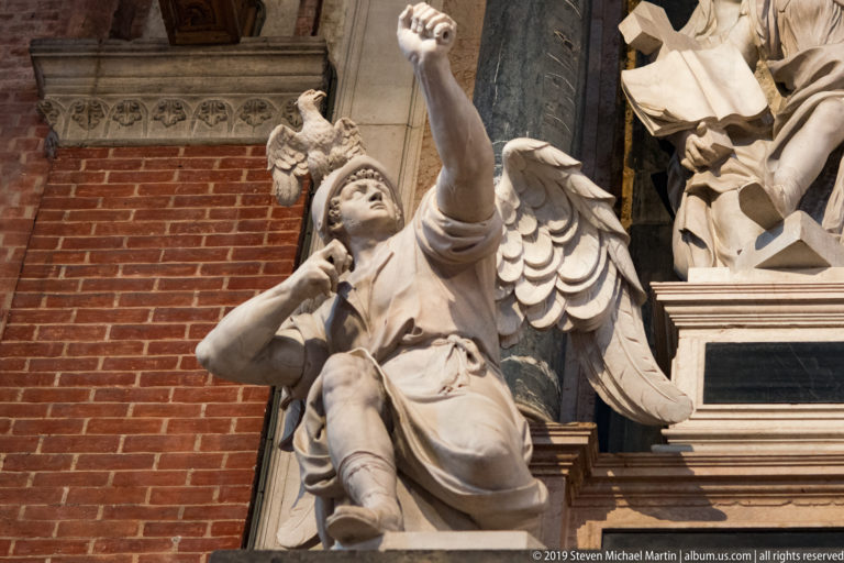 White marble angel with spread wings raises one arm and holds a trumpet to its mouth beside brickwork and a dark column.