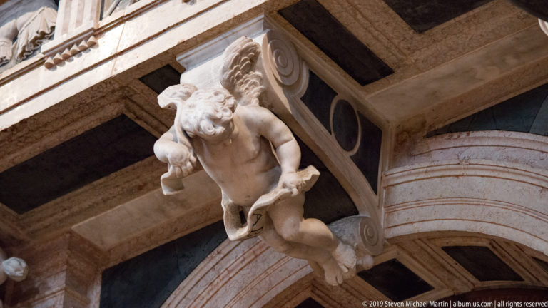 Carved winged putto projects outward beside a large volute and banded architectural element, viewed from below.