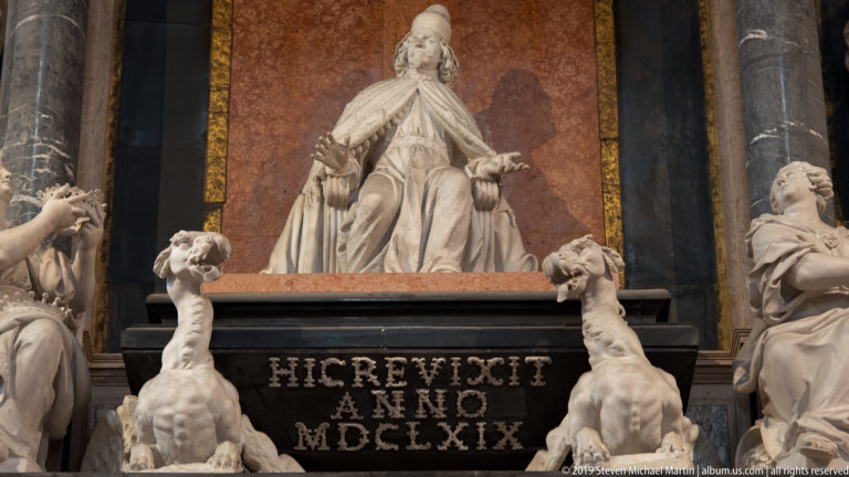 Seated white marble female figure centered above a black inscription panel bearing the Roman-numeral date MDCLXIX, with smaller sculptures below.