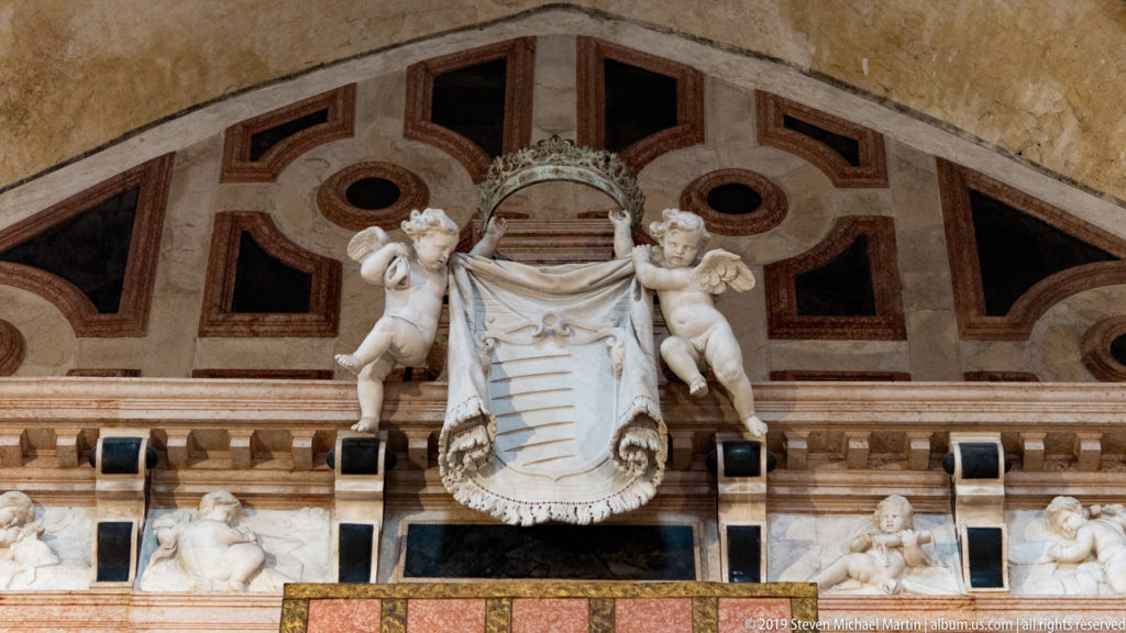 Two carved putti hold a striped heraldic shield beneath a crown above an architectural monument with geometric stone inlay.