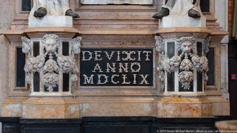 Black memorial panel reading “OBIIT ANNO MDCLXIX” between pale carved supports decorated with masks, fruit, leaves, and floral swags.