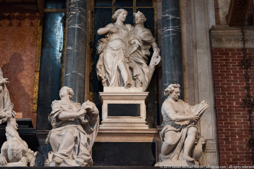 Central standing marble pair on a pedestal, flanked below by seated figures including one reading an open book, within a columned monument.