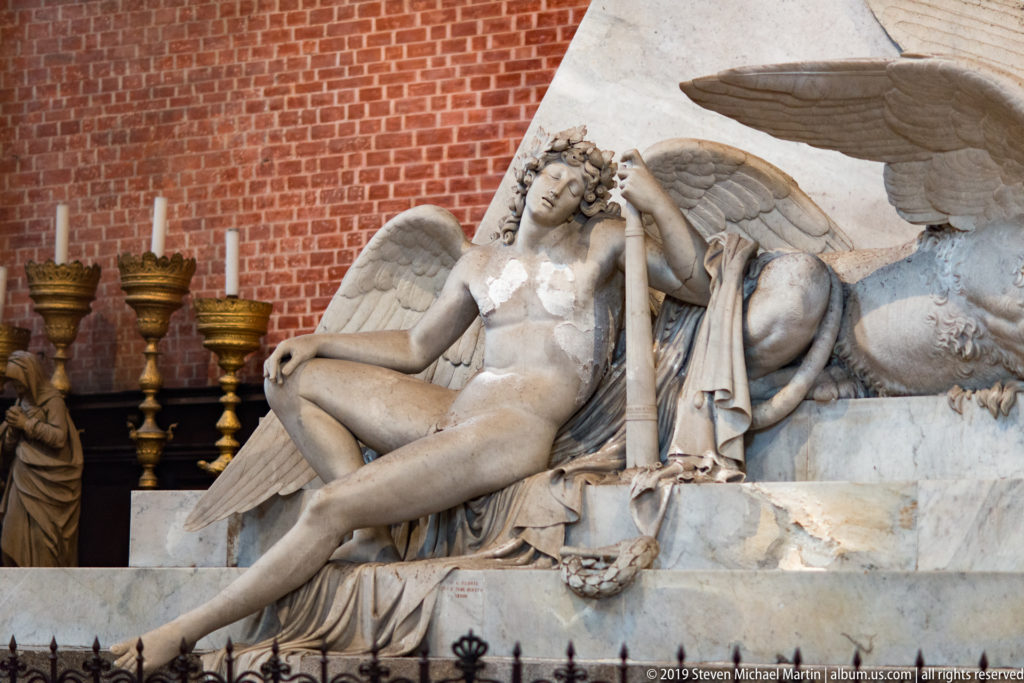 Reclining winged marble figure rests its head on one hand, with large feathered wings and drapery against a red-brick background.
