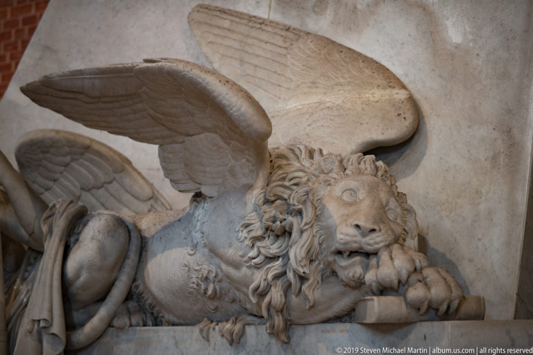 Recumbent winged marble lion with raised feathered wings, carved mane, and forepaws resting over a rounded form.