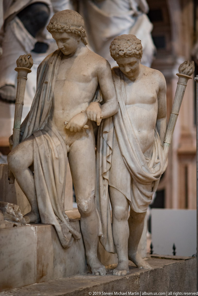 Two muscular marble male figures in partial drapery bend and brace beside columns as supporting figures within a larger monument.