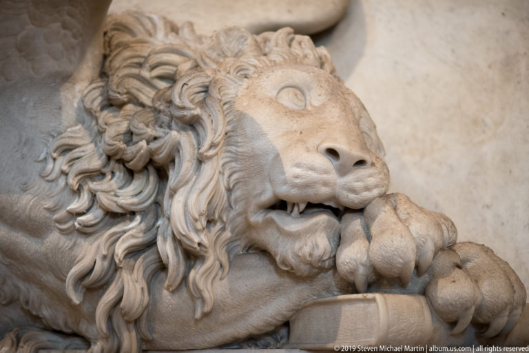 Close view of a carved marble lion with a thick curling mane and both forepaws resting on a rounded support.