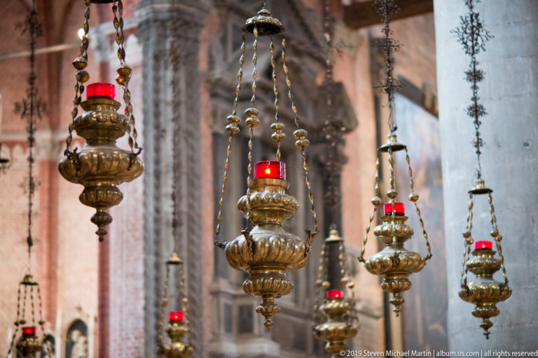 Multiple ornate brass hanging lamps on chains, several glowing red, suspended at varying depths inside the brick-and-stone church interior.