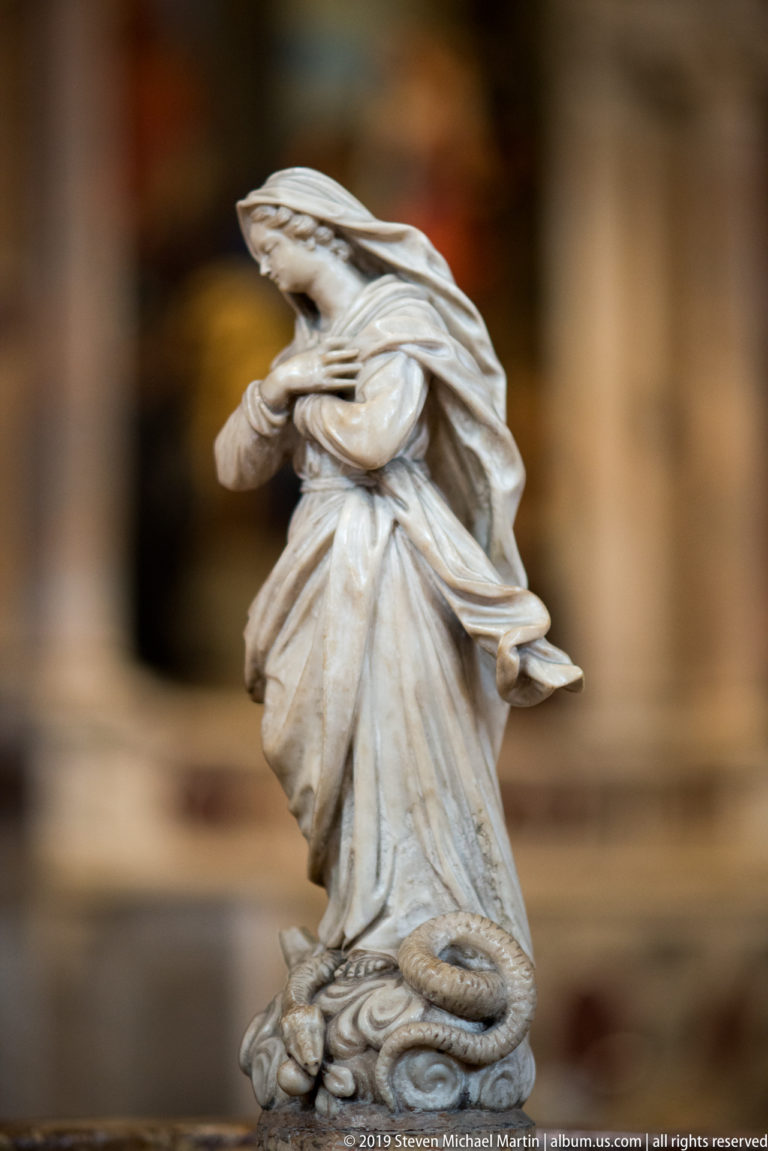 Small white marble figure of a kneeling woman leaning forward in deeply folded drapery, isolated against a blurred church interior.