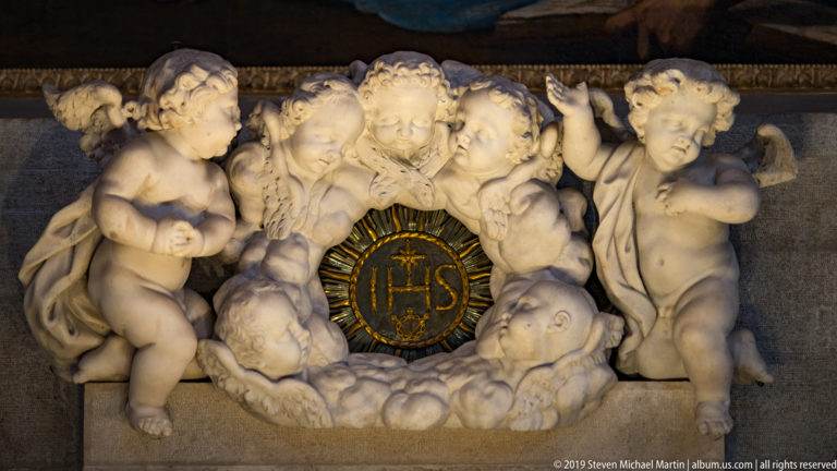 White marble putti crowd around a black circular medallion bearing a gold radiant IHS monogram.