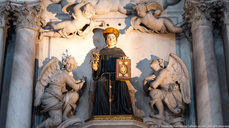 Dark-robed standing saint holding a decorated book, flanked by two pale kneeling angels beneath sculpted clouds.