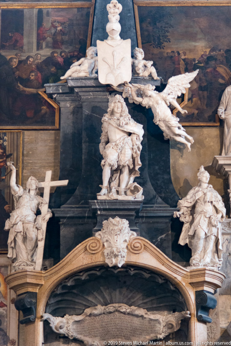 White marble winged figure stands before a dark central memorial, with heraldic ornament and additional sculpted figures arranged on both sides.