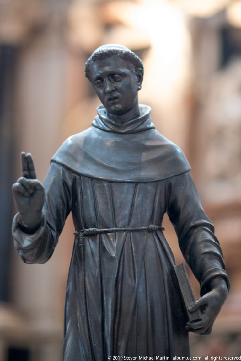 Dark metal statue of a Franciscan friar in a corded habit, raising his right hand in blessing and holding an object in his left.