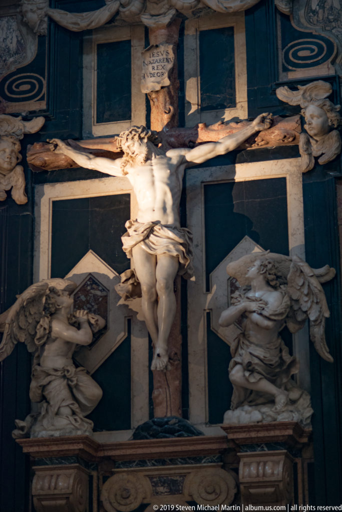 Crucified Christ centered on a dark funerary monument, with pale stone mourning figures and carved ornament arranged below and around the cross.