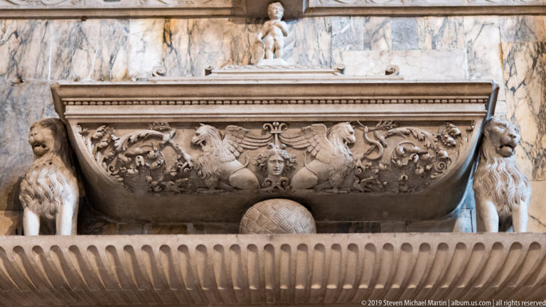 Carved stone tomb chest with scrolling foliage, central female mask, heraldic shield, and standing figures against veined marble.