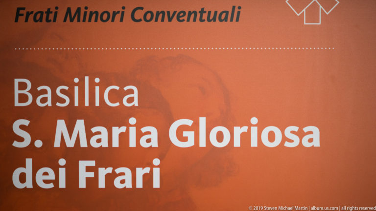 Orange sign reading “Basilica S. Maria Gloriosa dei Frari” in large white letters, with a faint religious image behind the text.