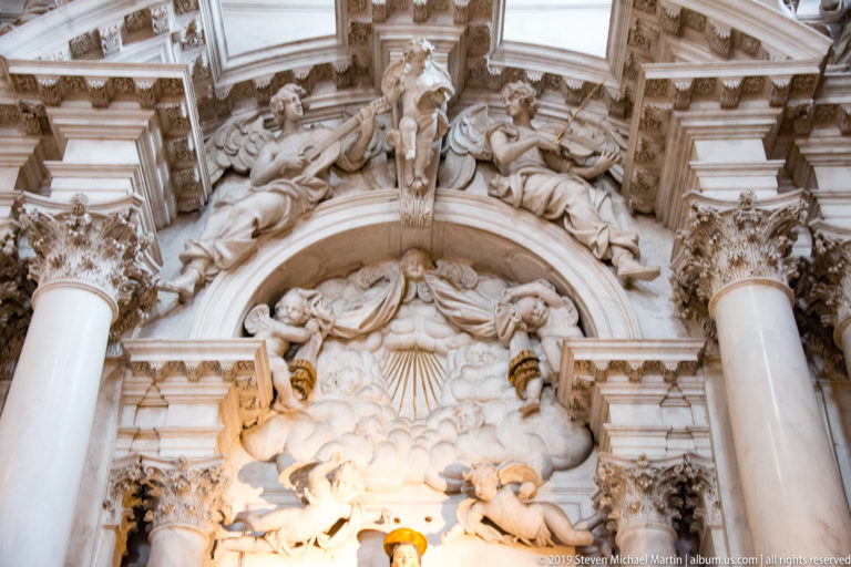 Upper section of an ornate stone altar with columns, angels, clouds, and a radiant dove-like emblem at the center.