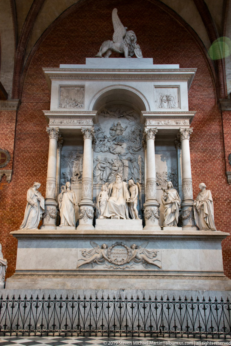 Large pale stone funerary monument against red brick, with paired columns, a central relief, seated figures, and a reclining figure above.