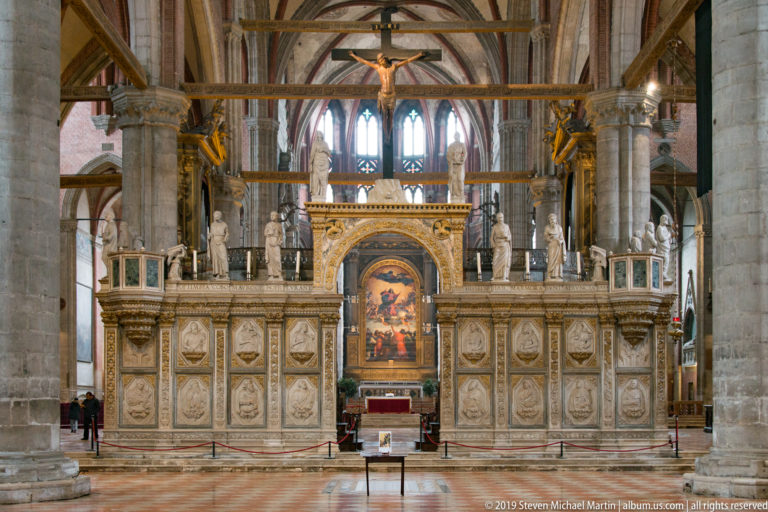 Large marble choir screen with relief panels, standing statues, central arched opening, and a crucifix above, aligned with the distant altar.
