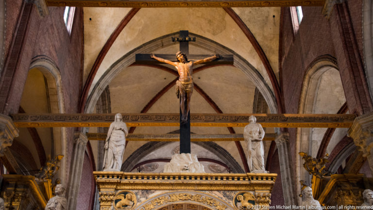 Large Crucifix suspended above the choir screen, with two standing statues below and Gothic vaulting and brick ribs behind.