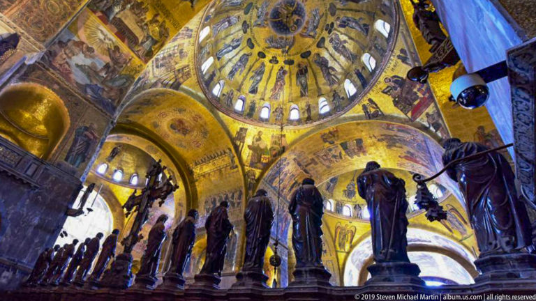 Dark standing sculptures silhouetted beneath luminous gold mosaic domes and arches inside St. Mark’s Basilica.