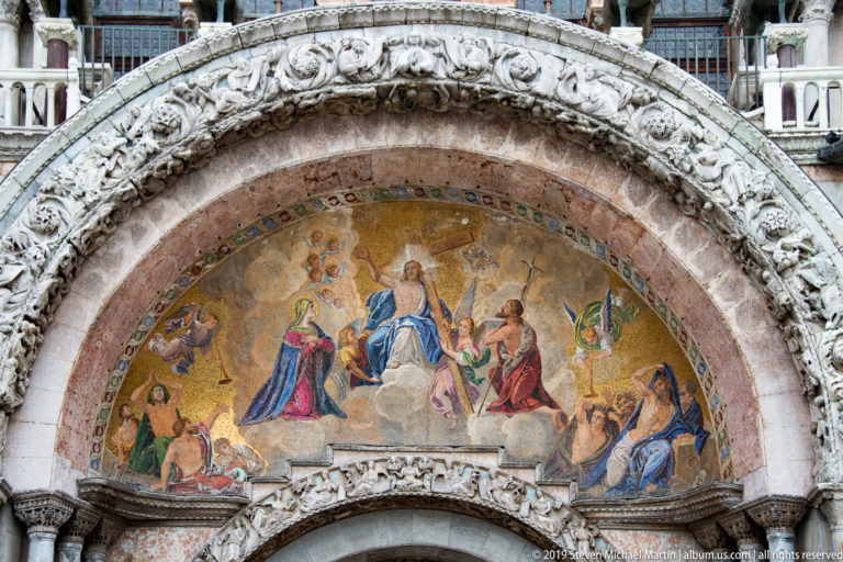 Semicircular gold mosaic with Christ and surrounding figures framed by reddish marble and carved stone arches.