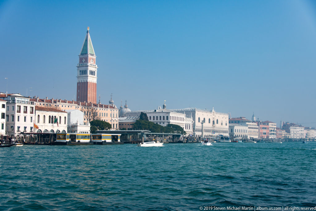 St. Mark’s Campanile above Venice’s waterfront, with vaporetto stops, boats, and lagoon water in the foreground.