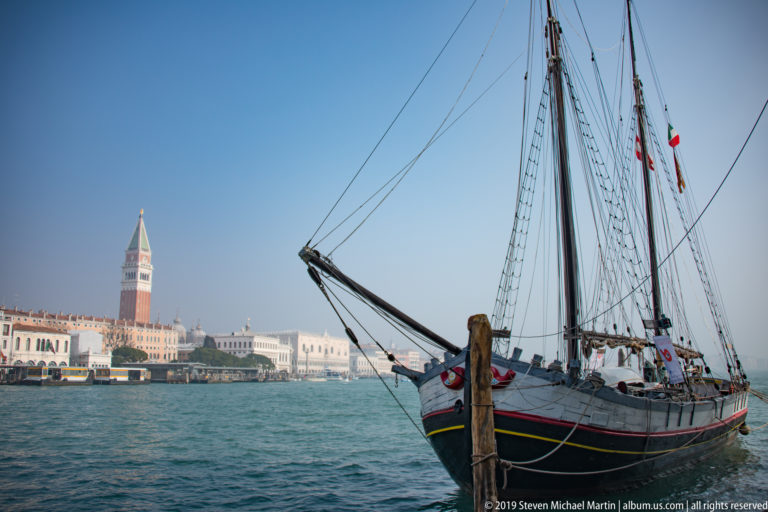 Rigged wooden sailing vessel on the Venetian lagoon with St. Mark’s Campanile and waterfront buildings in the background.