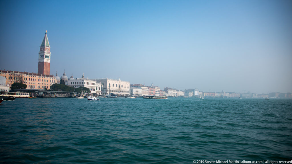St. Mark’s Campanile and waterfront buildings seen across blue-green lagoon water with small boats in the distance.
