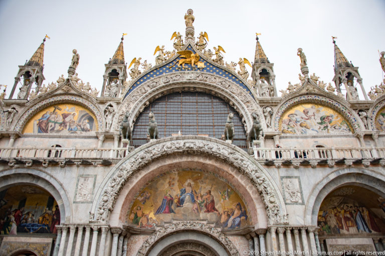 Upper façade of St. Mark’s Basilica with mosaic lunettes, central arched window, marble arcades, pinnacles, and statues.