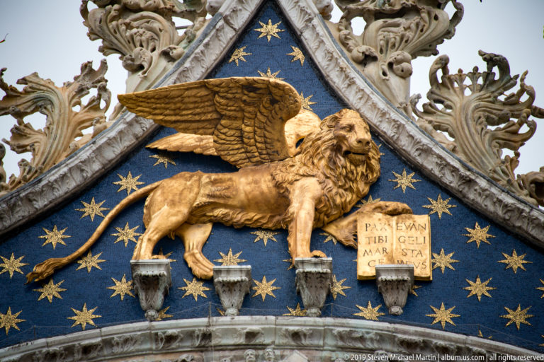 Gilded winged lion with open book on a blue star-patterned gable framed by ornate carved stonework.