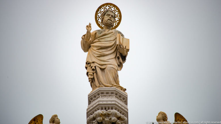 Haloed robed saint raising one hand and holding a book atop the roofline of St. Mark’s Basilica.