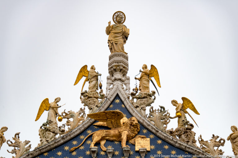 Blue star-patterned pediment with gilded winged lion, angels, and a haloed rooftop saint on St. Mark’s Basilica.