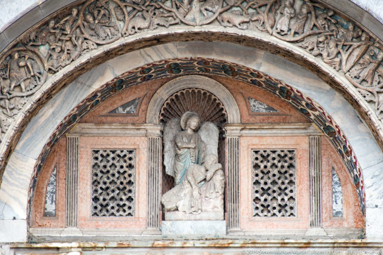 Robed figure beneath a shell niche between two pierced geometric panels in a semicircular basilica portal lunette.