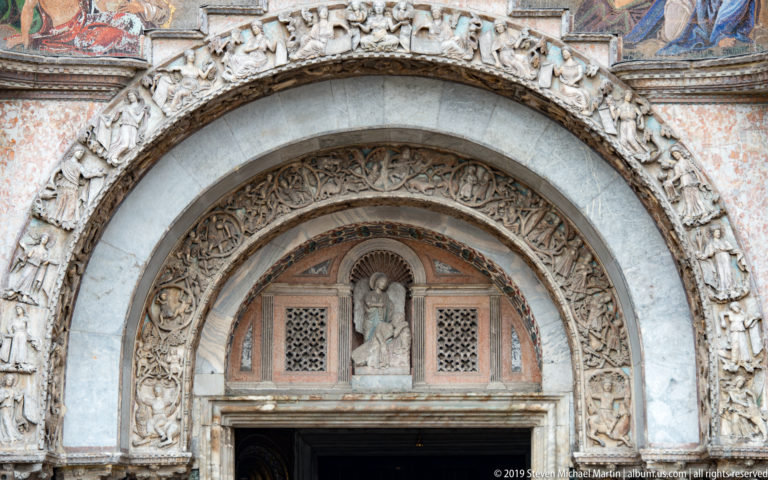 Concentric carved arches framing a small sculpted lunette and patterned grilles above a St. Mark’s Basilica doorway.