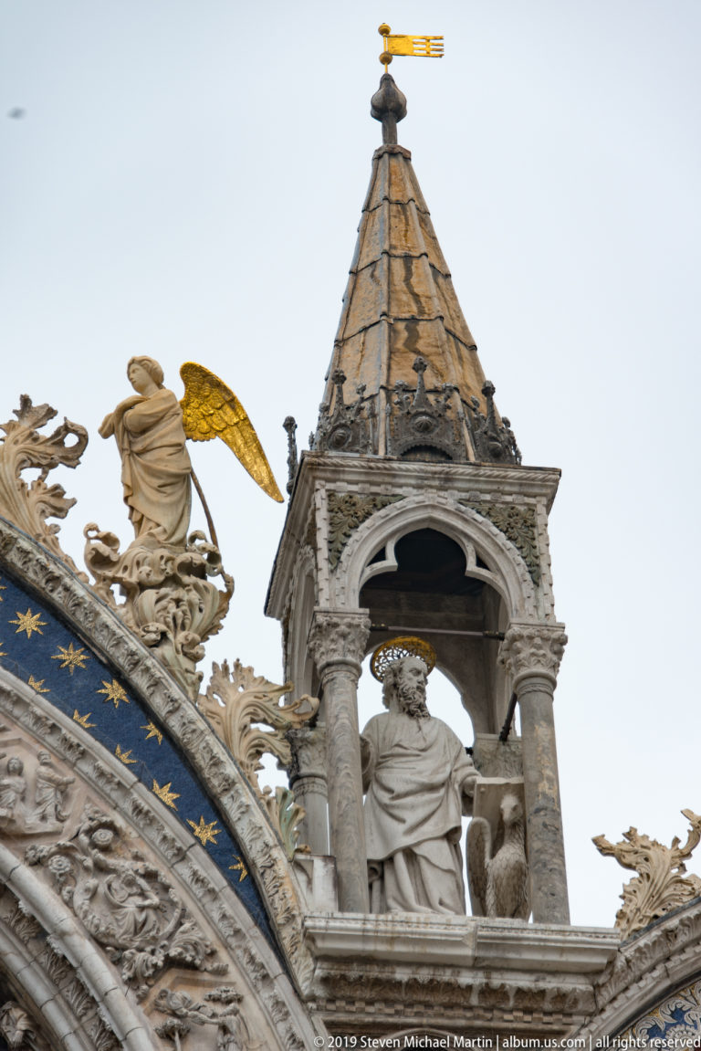 Small roof turret with standing saint beside a gilded angel and ornate cresting on St. Mark’s Basilica.