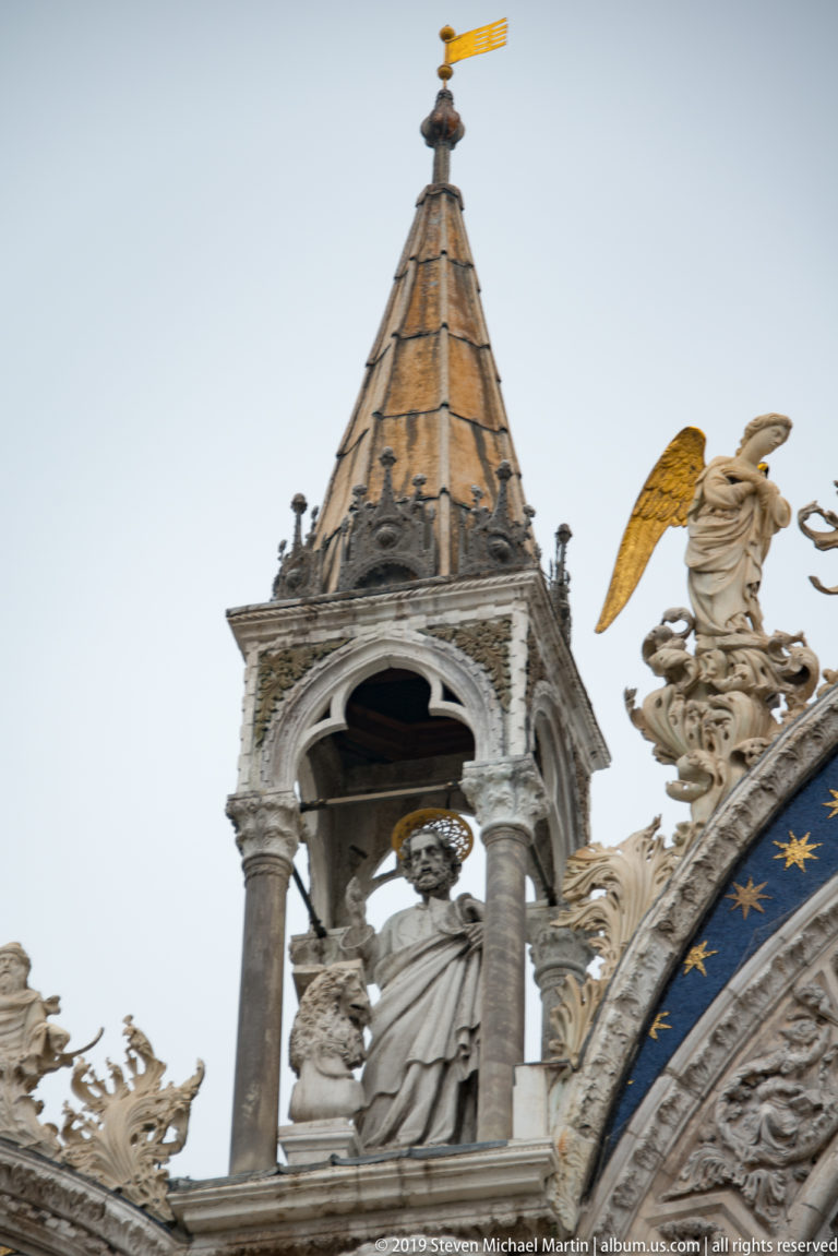 Roofline canopy with a robed statue surrounded by carved cresting and gilded-winged angels on St. Mark’s Basilica.