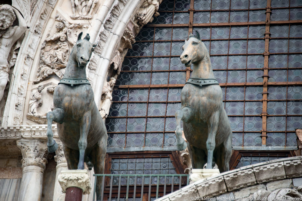 Two green-patinated bronze horse sculptures standing before a large gridded window on St. Mark’s Basilica.