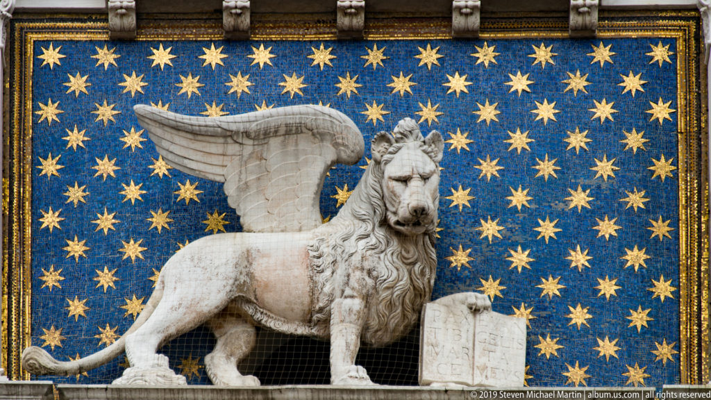 Winged Lion of St. Mark standing before a blue field filled with gold stars, beside an open book.