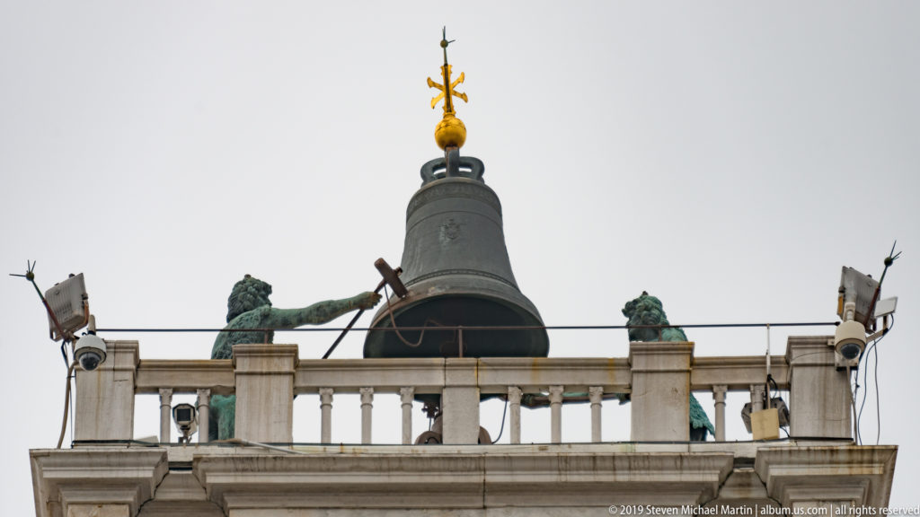 Large rooftop bell flanked by two bronze figures on St. Mark’s Clock Tower beneath a gilded finial.