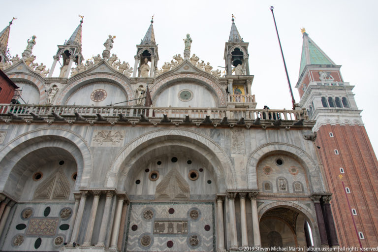Marble side façade of St. Mark’s Basilica beside the red-brick Campanile under an overcast sky.