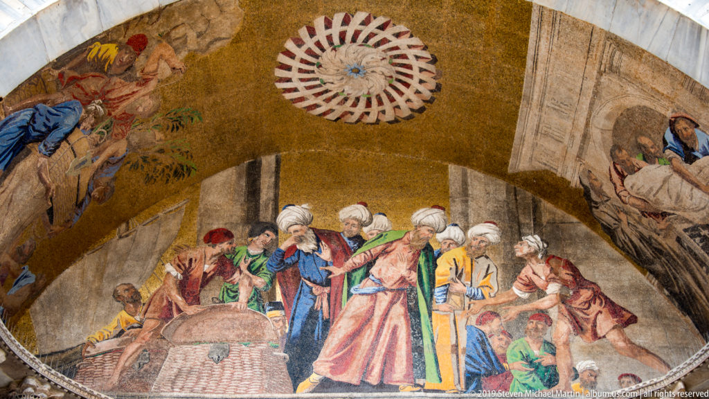 Gold mosaic lunette with turbaned figures gathered around a central object, framed by stone architecture and a rosette.