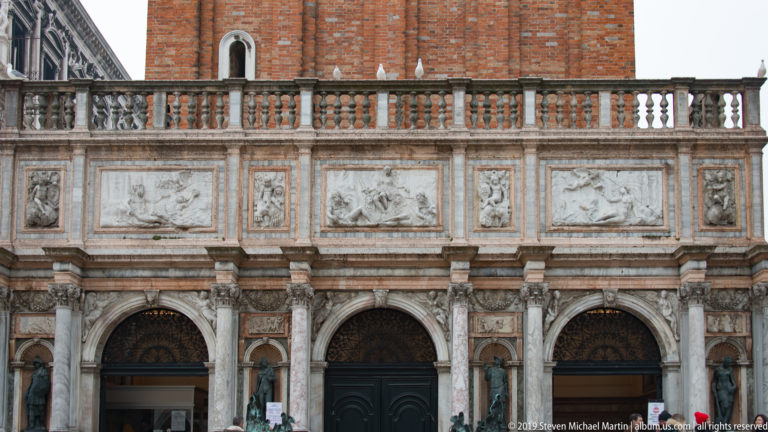 Three arched openings beneath a band of carved relief panels and balustrade on an ornate Venetian façade.