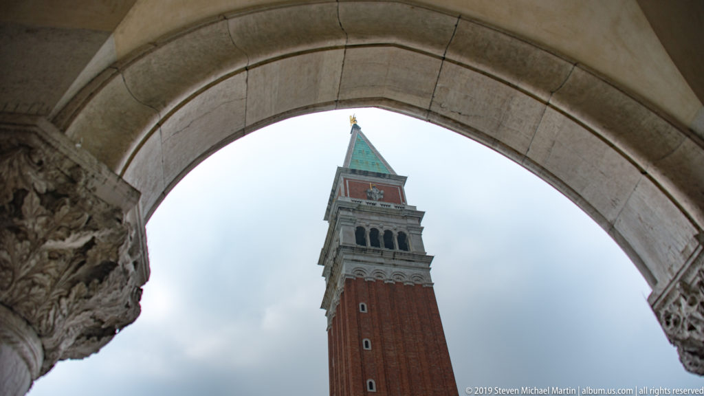 St. Mark’s Campanile seen through a broad stone arch, with its brick shaft and green roof against cloudy sky.