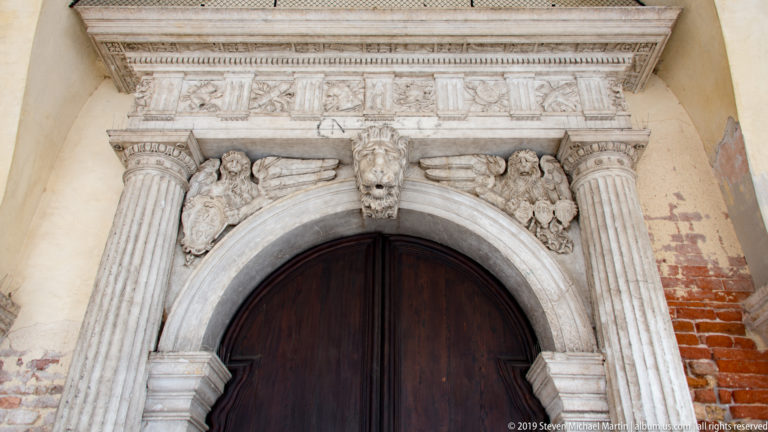 Arched wooden doorway framed by fluted stone pilasters, carved figures, entablature, and a central mask.