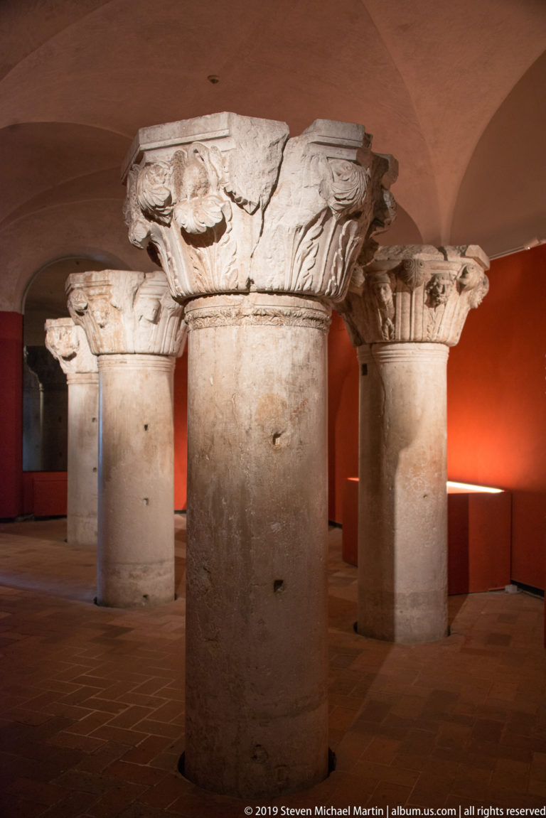 Four freestanding stone columns with weathered carved capitals displayed in a red-walled museum gallery.