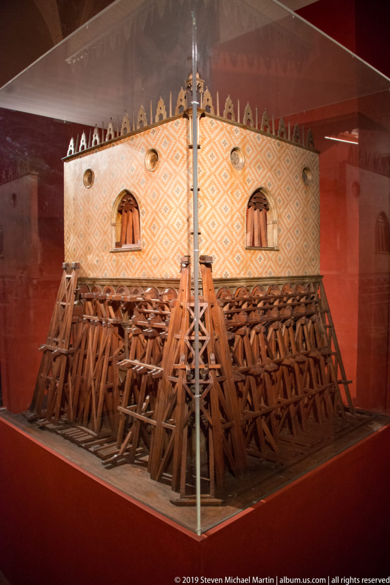 Large wooden architectural model with patterned upper walls and dense exposed bracing, displayed behind glass.