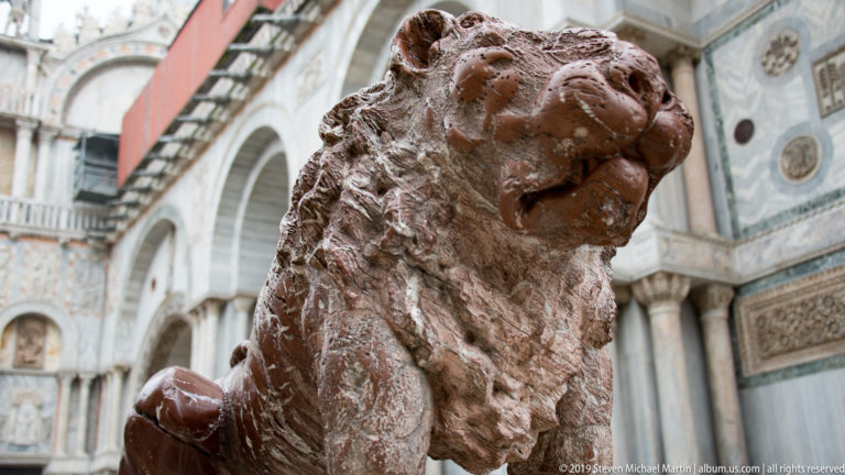 Close view of a weathered stone lion sculpture with St. Mark’s Basilica marble façade blurred behind it.